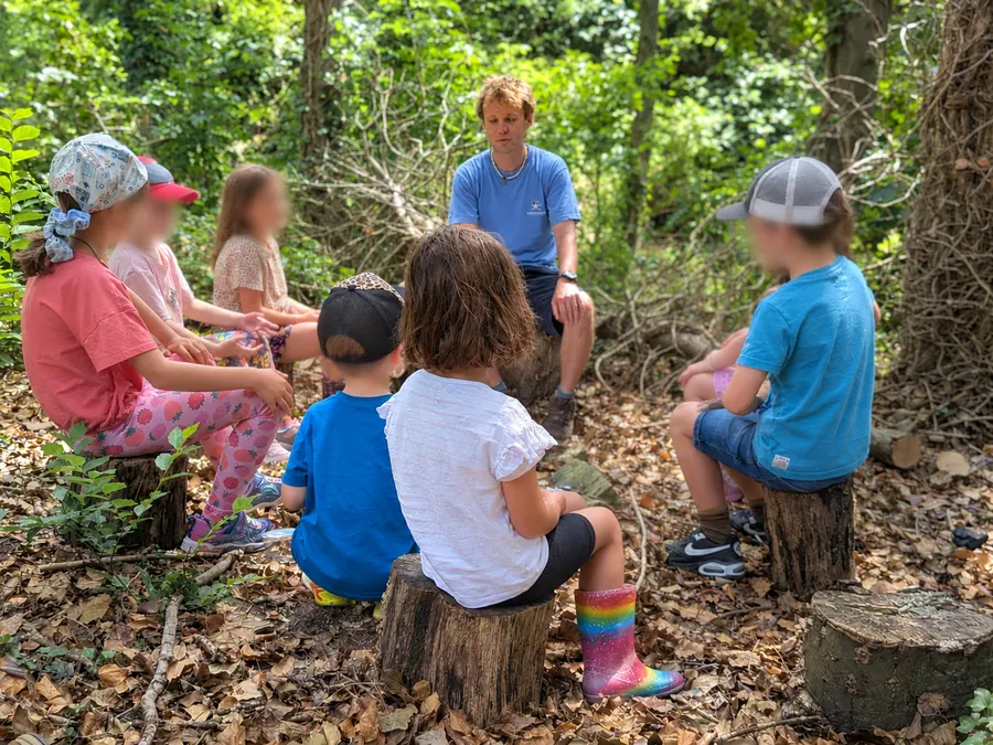 Ross sitting with a circle of young children on log stools in the woods
